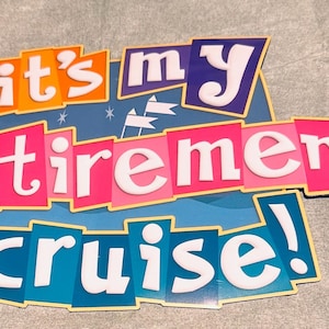 May include: A decorative sign featuring the text "it's my retirement cruise!" in white letters. The words are arranged on colorful, overlapping rectangular panels in shades of orange, purple, pink, and blue, with small white flag icons.