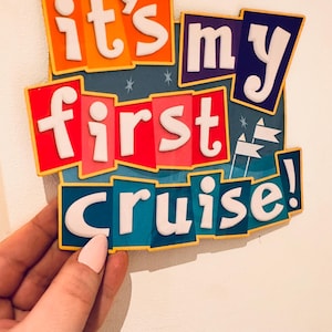 May include: A rectangular sign featuring the text "it's my first cruise!" in raised white lettering. The background consists of layered, colorful panels in orange, dark blue, red, and teal, accented with small white star and flag icons.