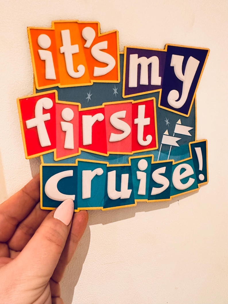 May include: A rectangular sign featuring the text "it's my first cruise!" in raised white lettering. The background consists of layered, colorful panels in orange, dark blue, red, and teal, accented with small white star and flag icons.