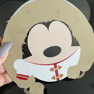 K&ouml;nnte beinhalten: Ein runder Aufkleber mit einer stilisierten Zeichentrickfigur mit schwarzen Ohren, die eine beige Kapuze mit Fellbesatz, eine wei&szlig;e Jacke mit rotem Saum und braune Knebelverschl&uuml;sse tr&auml;gt. Das Design hat eine flache, grafische &Auml;sthetik.