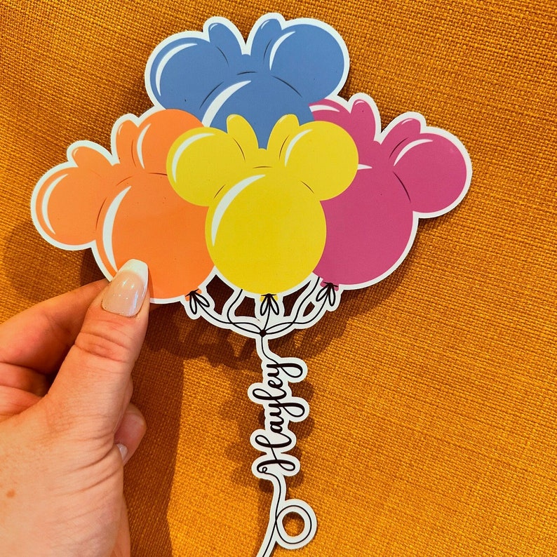 Pode incluir: Um conjunto de cinco bal&otilde;es de papel coloridos com orelhas de Mickey Mouse. Os bal&otilde;es s&atilde;o azuis, laranja, amarelos, rosa e roxos. Os bal&otilde;es est&atilde;o amarrados com um cord&atilde;o que diz "Hawley".