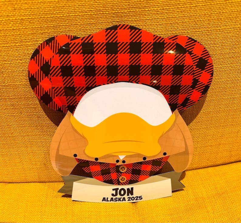 K&ouml;nnte beinhalten: Ein flaches, dekoratives Ornament in Form eines Entenkopfes mit einer rot-schwarz karierten M&uuml;tze. Die Ente hat einen wei&szlig;en Kopf, einen gelben Schnabel und einen beigen Hals. Ein Banner am unteren Rand zeigt den Text JON ALASKA 2025.