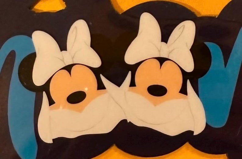 Op de afbeelding: Een decoratief wandbord met twee cartoon-muizenkoppen met grote witte strikken, perzikkleurige gezichten en zwarte oren. Het ontwerp is geplaatst tegen een donkere achtergrond met helderblauwe accenten en een levendige gele rand.