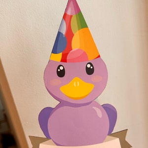 May include: A flat, purple duck-shaped cutout featuring a colorful polka-dot party hat topped with a small red star. The duck has a yellow beak and black eyes. A beige banner across the bottom displays the name ELLA in bold black capital letters.