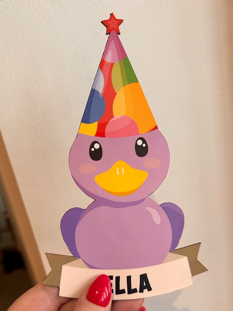 May include: A flat, purple duck-shaped cutout featuring a colorful polka-dot party hat topped with a small red star. The duck has a yellow beak and black eyes. A beige banner across the bottom displays the name ELLA in bold black capital letters.
