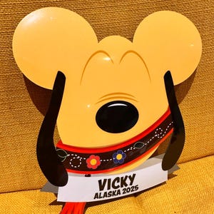 K&ouml;nnte beinhalten: Ein gelbes, ausgestanztes Schild in Form eines Cartoon-Hundekopfes mit langen schwarzen Ohren. Es zeigt ein rotes Halsband mit Blumenmuster und ein wei&szlig;es Banner am unteren Rand mit dem schwarzen Text VICKY ALASKA 2025 sowie roten Quasten.