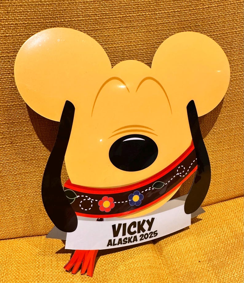 K&ouml;nnte beinhalten: Ein gelbes, ausgestanztes Schild in Form eines Cartoon-Hundekopfes mit langen schwarzen Ohren. Es zeigt ein rotes Halsband mit Blumenmuster und ein wei&szlig;es Banner am unteren Rand mit dem schwarzen Text VICKY ALASKA 2025 sowie roten Quasten.