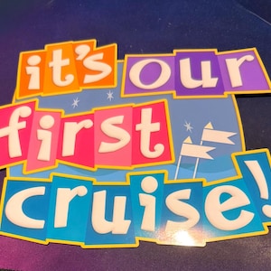 May include: A rectangular sign with a blue background featuring white star and flag icons. The text reads "it's our first cruise!" in white, bubbly, lowercase letters set against overlapping orange, purple, pink, and blue rectangular blocks.
