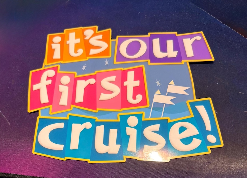 May include: A rectangular sign with a blue background featuring white star and flag icons. The text reads "it's our first cruise!" in white, bubbly, lowercase letters set against overlapping orange, purple, pink, and blue rectangular blocks.