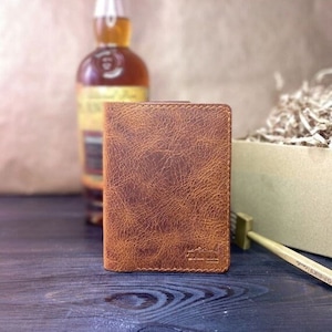 Puede incluir: Una cartera de cuero marrón con una superficie texturizada. La cartera tiene un pequeño logotipo en la parte delantera.
