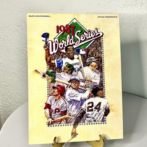 May include: A vintage 1987 World Series official program featuring illustrations of baseball players. The cover has the text "1987 World Series" and "Major League Baseball". The program is displayed on a gold-colored stand.