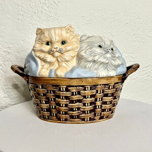 May include: A ceramic casserole dish shaped like a wicker basket with two kittens inside. The kittens are made of ceramic and are painted in shades of white, gray, and orange. The basket is painted brown and has a blue and white blanket inside.