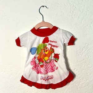 May include: A white t-shirt with red trim and a red ruffle hem. The shirt features a cartoon character holding a gift box with the text "Fun's a poppin'" and "Popples".