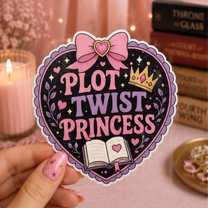 May include: A heart-shaped sticker with a black background featuring the pink text "PLOT TWIST PRINCESS". The design includes a light pink bow at the top, a gold crown with a pink gem, a small open book with a heart, and decorative floral vines.