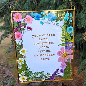 May include: A gold framed glass picture with pressed flowers and greenery around the edges. The centre of the picture has a white background with the text "your custom text, scripture, poem, lyrics, or message here" printed in brown.