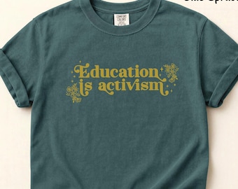 Puede incluir: Una camiseta de manga corta color abeto azul con cuello redondo y mangas remangadas. El frente presenta el texto dorado "Education is activism" en una fuente estilizada, acentuado con pequeñas ilustraciones florales y estrellas a los lados.
