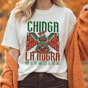 May include: Off-white t-shirt featuring a colorful eagle design with the text "CHINGA LA MIGRA" in green and red. The eagle has orange, teal, and red wings. The shirt is paired with brown pants and an orange sweater.