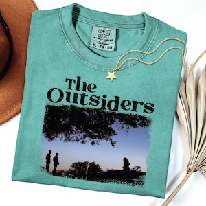 May include: Teal-colored t-shirt with the text "The Outsiders" and a graphic of silhouetted figures under a tree. A gold star pendant necklace and a brown hat are next to the shirt. The shirt is size XL.