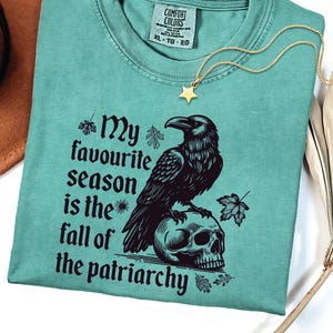 May include: Light green t-shirt with the text "My favourite season is the fall of the patriarchy" and a graphic of a raven perched on a skull. A gold star pendant necklace is draped on the shirt. A brown hat is in the background.