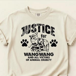 May include: A cream-colored short-sleeve t-shirt featuring a black graphic of a dog with puppies, two paw prints, and the text: JUSTICE for WANGWANG AND ALL VICTIMS OF ANIMAL CRUELTY. The shirt has a crew neckline and a Comfort Colors brand label.