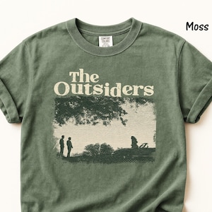 May include: A moss green short-sleeve t-shirt featuring a graphic print of the text The Outsiders above a scene with two figures standing near a car under a tree. The shirt has a crew neckline and a label inside the collar reading Comfort Colors.