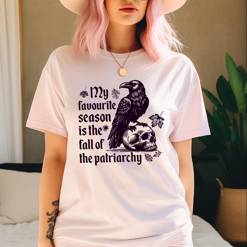 May include: White t-shirt featuring a black raven perched on a skull, with the text "My favourite season is the fall of the patriarchy." The design includes leaf accents. A casual, graphic tee.