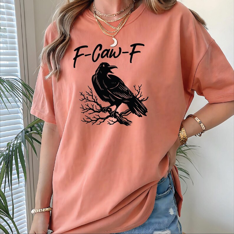 The image shows a woman wearing a relaxed-fit, peach-toned t-shirt featuring a bold black graphic. The design showcases a detailed raven perched on a bare tree branch, with the words “F-Caw-F” written above in a playful, stylized font. The model is accessorized with layered gold necklaces, including a crescent moon pendant, a watch, and bracelets, giving the outfit a chic and edgy vibe. She's standing indoors with houseplants in the background, adding a casual, natural atmosphere.