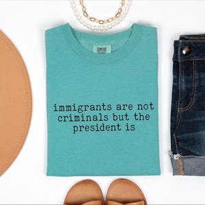 May include: A turquoise t-shirt with the text "immigrants are not criminals but the president is" in black. The shirt is displayed with denim shorts, sandals, and a necklace.