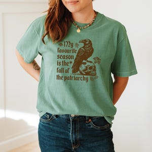 May include: A sage green t-shirt with a brown graphic of a raven perched on a skull. The text "My favourite season is the fall of the patriarchy" is printed below the raven and skull.