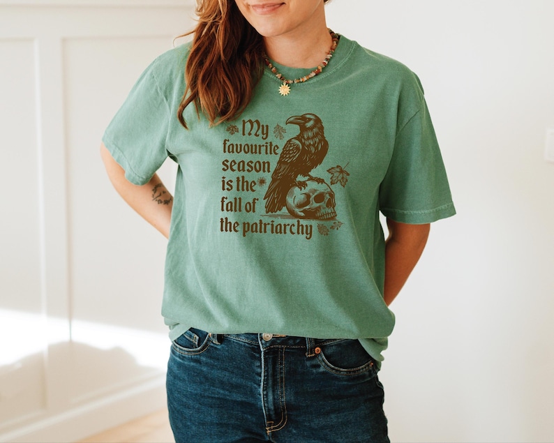 May include: A sage green t-shirt with a brown graphic of a raven perched on a skull. The text "My favourite season is the fall of the patriarchy" is printed below the raven and skull.