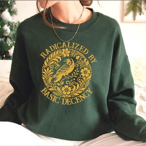 May include: Dark green sweatshirt with a gold graphic. The graphic features a raven inside a circular floral design with the words "RADICALIZED BY BASIC DECENCY". The sweatshirt is a crew neck style.