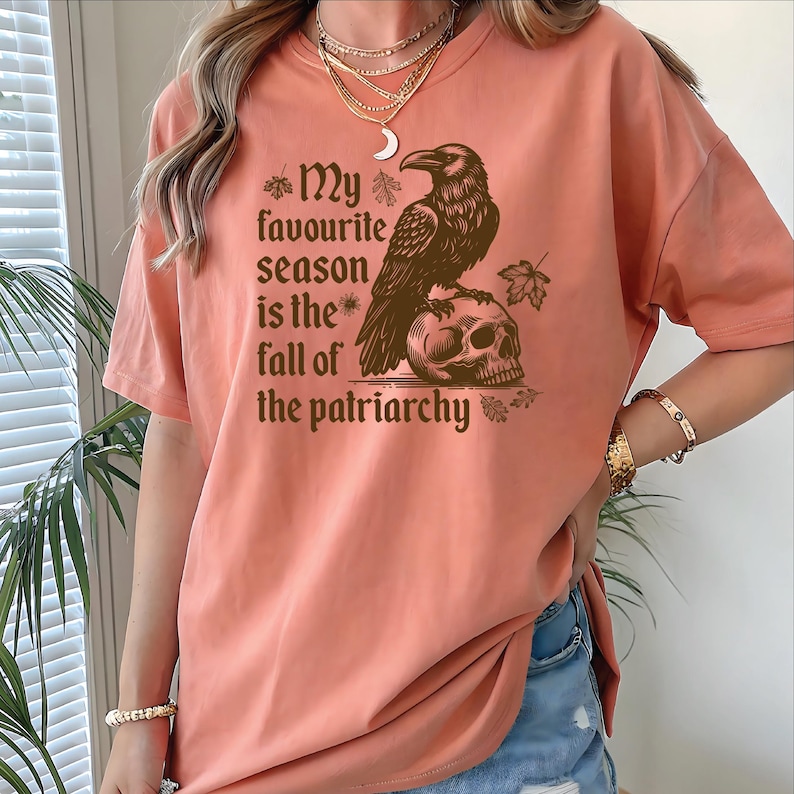 May include: Peach-colored t-shirt featuring a brown raven perched on a skull. The shirt has the text "My favourite season is the fall of the patriarchy" in a decorative font. The shirt is short-sleeved and has a relaxed fit.
