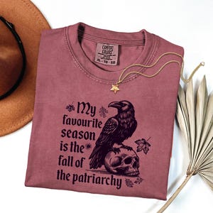 May include: A mauve t-shirt with the text "My favourite season is the fall of the patriarchy" and a graphic of a raven perched on a skull. A gold star pendant necklace and a brown hat are also in the image.