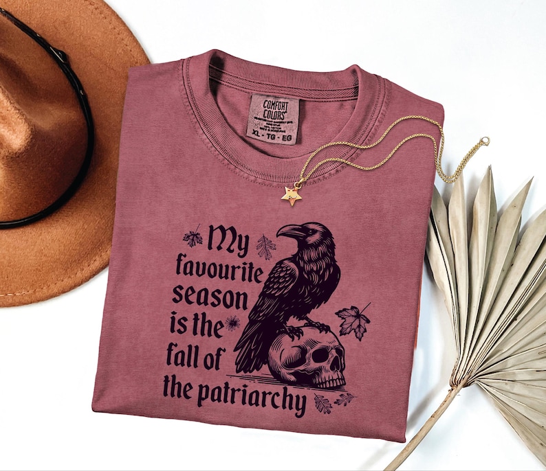 May include: A mauve t-shirt with the text "My favourite season is the fall of the patriarchy" and a graphic of a raven perched on a skull. A gold star pendant necklace and a brown hat are also in the image.