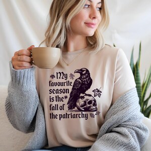 May include: Beige t-shirt with a black graphic of a raven perched on a skull, accompanied by the text "My favourite season is the fall of the patriarchy." The design includes leaf accents. The shirt is a casual wear item.