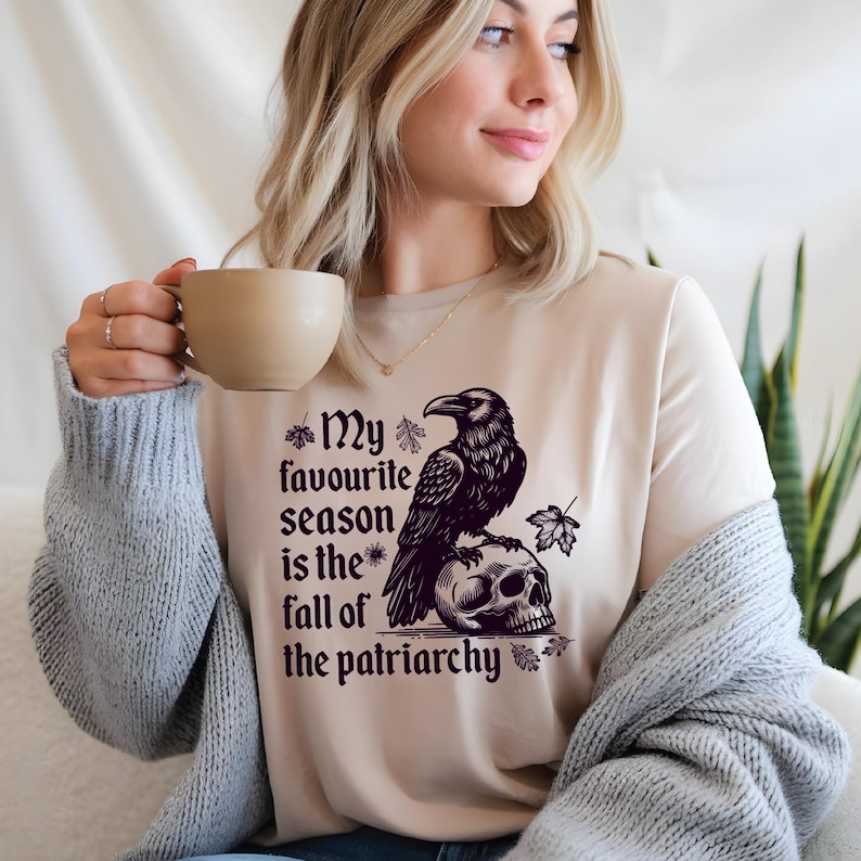 May include: Beige t-shirt with a black graphic of a raven perched on a skull, accompanied by the text "My favourite season is the fall of the patriarchy." The design includes leaf accents. The shirt is a casual wear item.