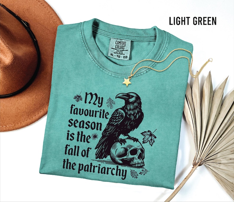 May include: Light green t-shirt with the text "My favourite season is the fall of the patriarchy" and a graphic of a raven perched on a skull. A gold star pendant necklace is draped on the shirt. A brown hat is in the background.