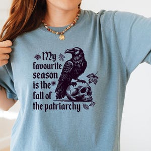 May include: A light blue t-shirt with a graphic featuring a black raven perched on a skull. The text reads "My favourite season is the fall of the patriarchy". The design includes autumn leaves.