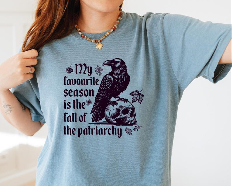 May include: A light blue t-shirt with a graphic featuring a black raven perched on a skull. The text reads "My favourite season is the fall of the patriarchy". The design includes autumn leaves.