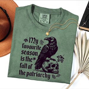 The image shows a folded sage green Comfort Colors T-shirt with a bold gothic-style design. The artwork features a large black raven perched on a human skull, surrounded by scattered autumn leaves. Next to the raven is the empowering phrase:

“My favourite season is the fall of the patriarchy.”

The shirt is styled with accessories around it: a gold star necklace, a black necklace with a cross, a brown wide-brim hat, and dried palm leaves, creating an earthy, rustic aesthetic.