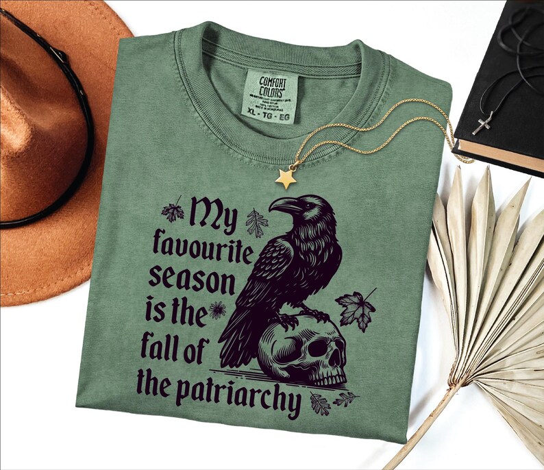 The image shows a folded sage green Comfort Colors T-shirt with a bold gothic-style design. The artwork features a large black raven perched on a human skull, surrounded by scattered autumn leaves. Next to the raven is the empowering phrase:

“My favourite season is the fall of the patriarchy.”

The shirt is styled with accessories around it: a gold star necklace, a black necklace with a cross, a brown wide-brim hat, and dried palm leaves, creating an earthy, rustic aesthetic.