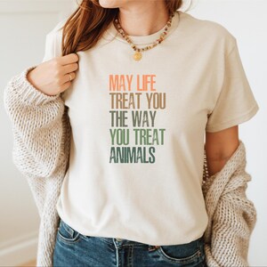 May include: A beige t-shirt with a message that reads "May life treat you the way you treat animals". The text is in a rainbow gradient.