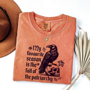 May include: A coral-colored Comfort Colors crew neck t-shirt with the text "My favourite season is the fall of the patriarchy" and a raven perched on a skull. A gold necklace and a brown hat are also visible.