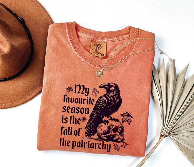 May include: A coral-colored Comfort Colors crew neck t-shirt with the text "My favourite season is the fall of the patriarchy" and a raven perched on a skull. A gold necklace and a brown hat are also visible.