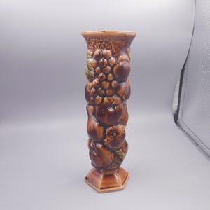 May include: A brown ceramic vase with a textured surface, featuring a relief design of various fruits. The vase has a hexagonal base and a cylindrical shape, with a detailed depiction of grapes, apples, and other fruits.