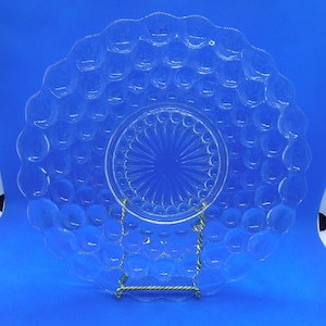 Puede incluir: Un plato de vidrio transparente con un patrón de burbujas en relieve alrededor del borde y un patrón de estrella estrellada más pequeño y concéntrico en el centro. El plato está sobre un soporte de metal dorado.