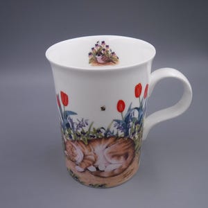 Pode incluir: Caneca de cerâmica branca com uma alça, apresentando uma ilustração colorida de um gato malhado laranja dormindo em um jardim com tulipas vermelhas, flores azuis e uma abelha. Uma pequena decoração floral está na borda.