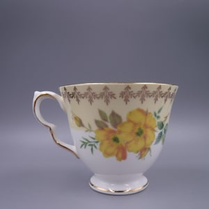 May include: A white teacup with a yellow floral design and gold trim. The cup features a decorative handle and a scalloped rim with a brown leaf pattern. The flowers are yellow with green leaves.