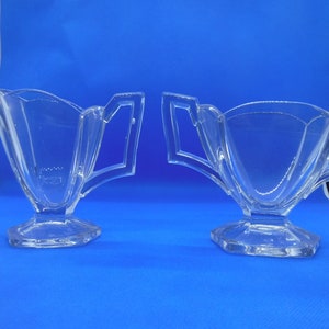May include: A set of two clear glass sugar bowls with a unique design. The bowls have a wide, flared base and a narrow, curved top. The handles are attached to the sides of the bowls and are shaped like a crescent moon.