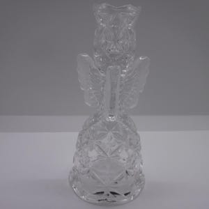 May include: A clear glass angel bell with a detailed, cut-glass design. The bell has a rounded base and a winged angel figure on top.